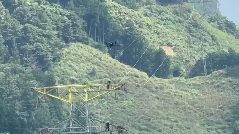山岳地の送電線工事で、ドローンが鉄塔間にパイロットロープを空中延線している様子