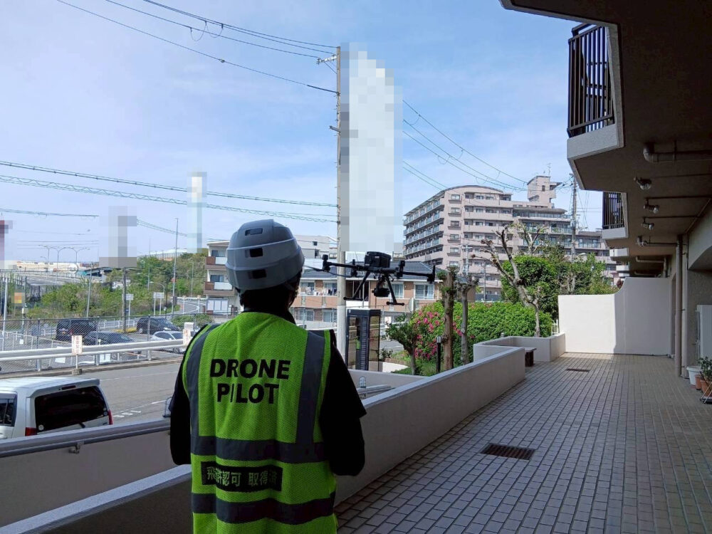 DRONE PILOTのベストを着た操縦者が、マンションの敷地内でドローンを飛行させている