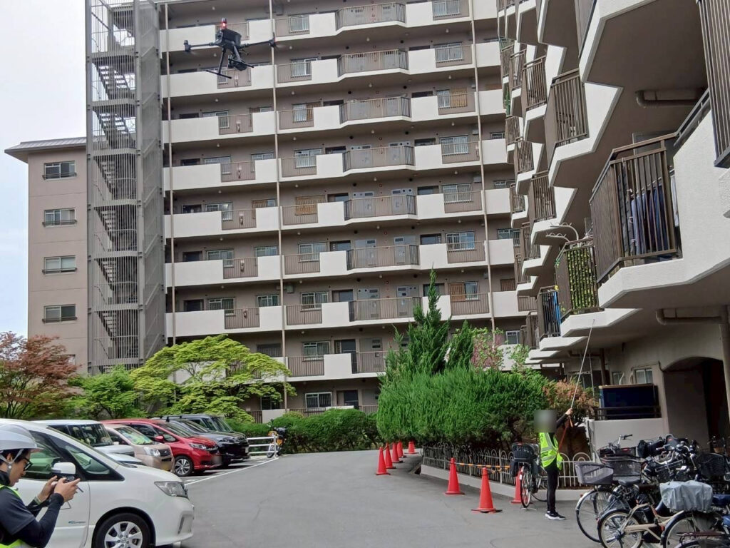 ドローンがマンションの外壁に沿って飛行し、地上ではカラーコーンで区画し補助者が立入管理を行っている