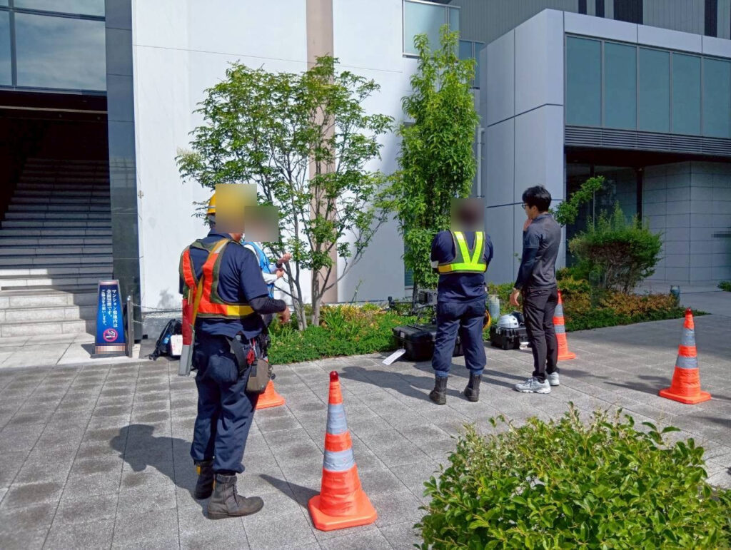 ドローン外壁調査の現場体制。安全ベストとヘルメットを着用した作業員が離着陸場所を管理し、機材を配置している