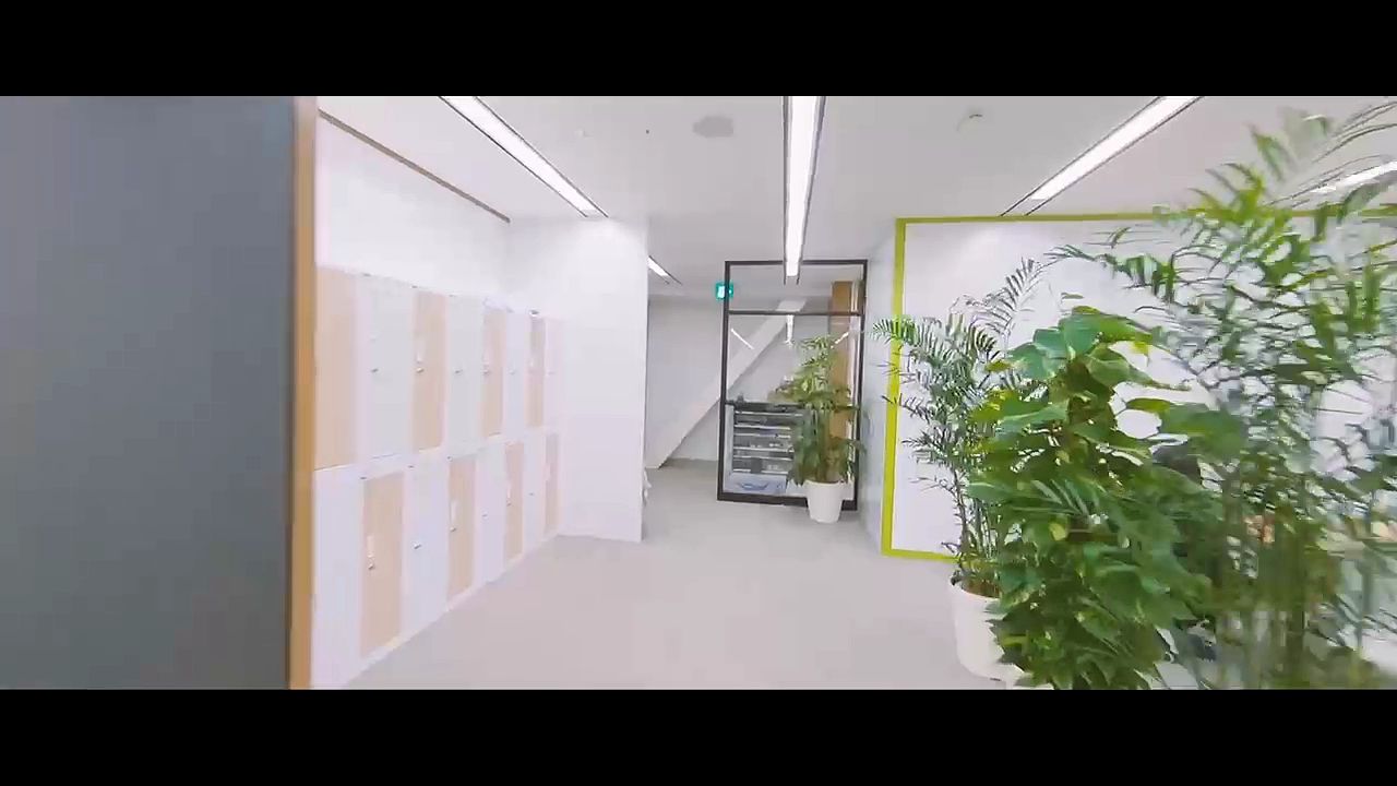 白いロッカーと観葉植物が並ぶ、明るいオフィスの通路。マイクロドローンで撮影した映像の一場面